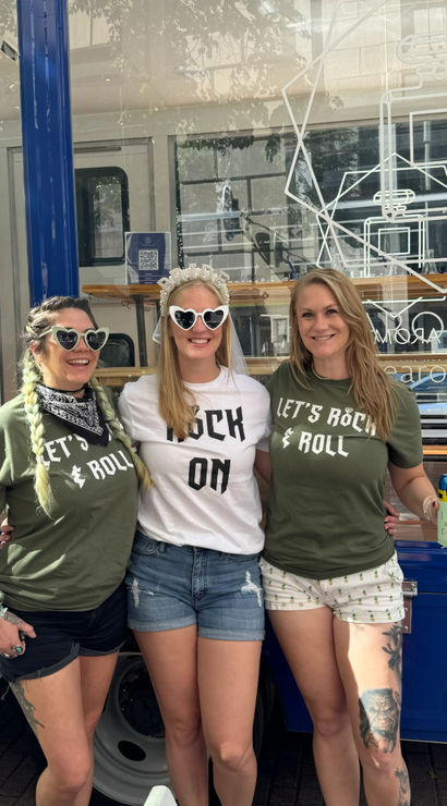 Three smiling women posing on a city sidewalk in front of a blue food truck — bachelorette in center with veil and heart sunglasses, friends in matching 'Let’s Rock & Roll' shirts.