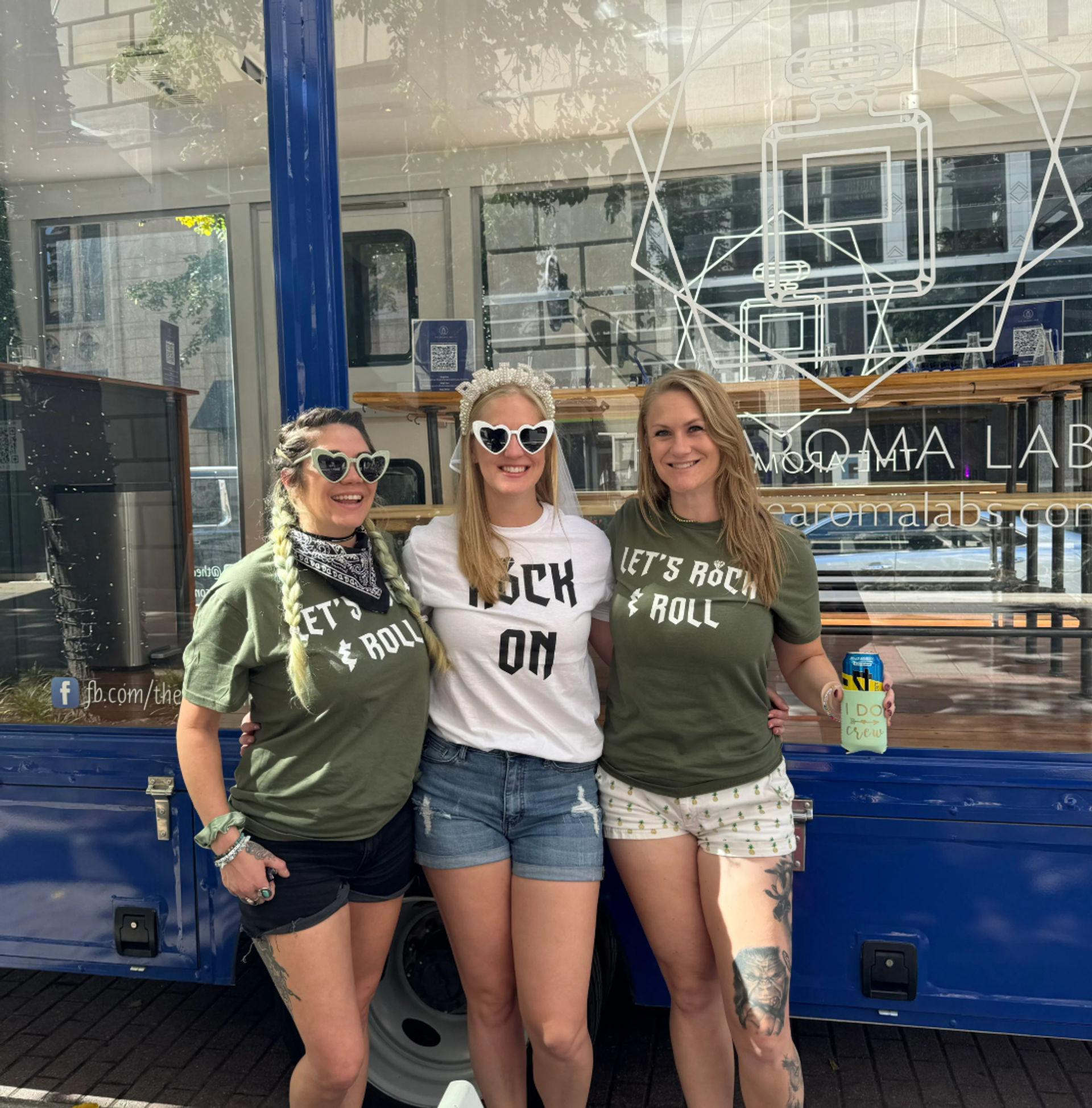 Three smiling women posing on a city sidewalk in front of a blue food truck — bachelorette in center with veil and heart sunglasses, friends in matching 'Let’s Rock & Roll' shirts.