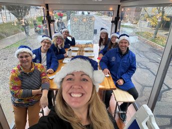 Smiling festive group in blue-and-white Santa hats taking a selfie inside a glass-sided holiday trolley with a wooden table, string lights and a QR code on the rear window, parked on a leafy city street.