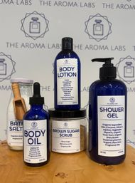 Blue-labeled natural body care set on a wooden counter: glass jar of bath salts with wooden scoop, blue dropper body oil, brown sugar scrub jar, body lotion bottle, and pump shower gel — home spa essentials.