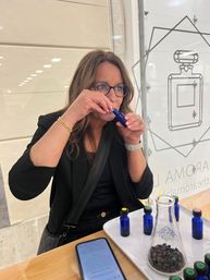 Woman in glasses sniffing a cobalt-blue perfume sample at a fragrance bar counter, tray of matching vials and a beaker of coffee beans on a wooden table with a smartphone nearby