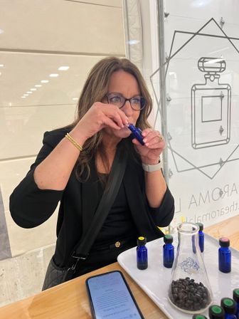 Woman in glasses sniffing a cobalt-blue perfume sample at a fragrance bar counter, tray of matching vials and a beaker of coffee beans on a wooden table with a smartphone nearby