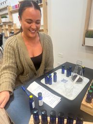 Smiling person mixing fragrances at a DIY scent bar — cobalt-blue sample bottles, glass beaker, pipette and handwritten notes on a tray in a bright modern studio.