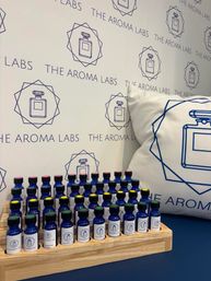 Blue glass fragrance sample vials with colorful caps neatly arranged on a wooden tray in a scent-bar display, set against patterned branded wallpaper and a matching perfume-logo pillow
