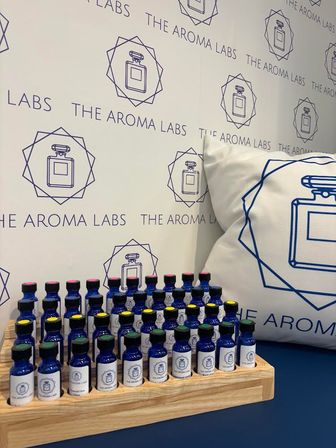 Blue glass fragrance sample vials with colorful caps neatly arranged on a wooden tray in a scent-bar display, set against patterned branded wallpaper and a matching perfume-logo pillow