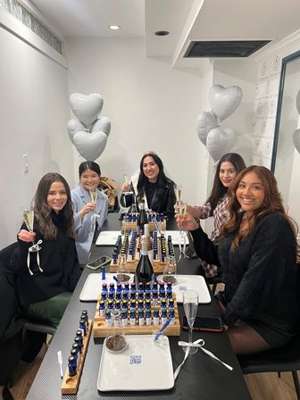 Five women toasting with champagne at an indoor scent-making party, trays of small fragrance bottles on the table, silver heart balloons and one guest wearing a tiara.