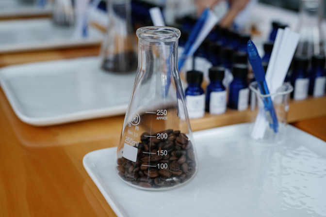 Erlenmeyer flask filled with coffee beans on a white tray, with blue sample bottles and pens blurred in the background, playful lab-style coffee display.