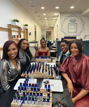 Five women smiling at a perfume workshop in a modern fragrance studio, gathered around a table filled with rows of small blue scent bottles, color-coded caps, droppers and glass beakers during a scent-blending class.