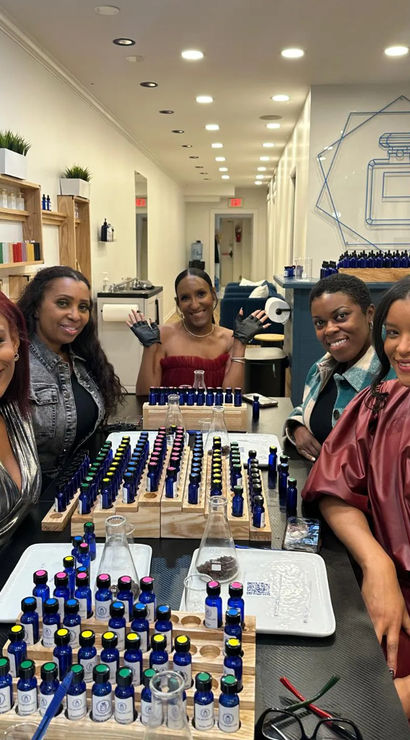 Five smiling women at a modern perfume-blending workshop in a boutique scent bar, arranging rows of cobalt sample bottles, pipettes and glass beakers.