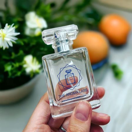 Hand holding a clear glass perfume bottle with a minimalist geometric logo, blurred white daisies and oranges in the background — bright home-fragrance lifestyle shot.