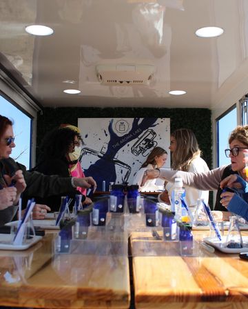 Sunny mobile pop-up fragrance workshop inside a trailer, people sampling scents from blue vials and droppers at a long wooden communal table.