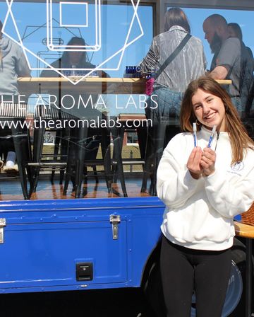 Smiling young woman holding two perfume sample vials outside a blue mobile fragrance pop-up truck with glass window seating and customers inside