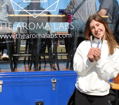 Smiling young woman holding two perfume sample vials outside a blue mobile fragrance pop-up truck with glass window seating and customers inside