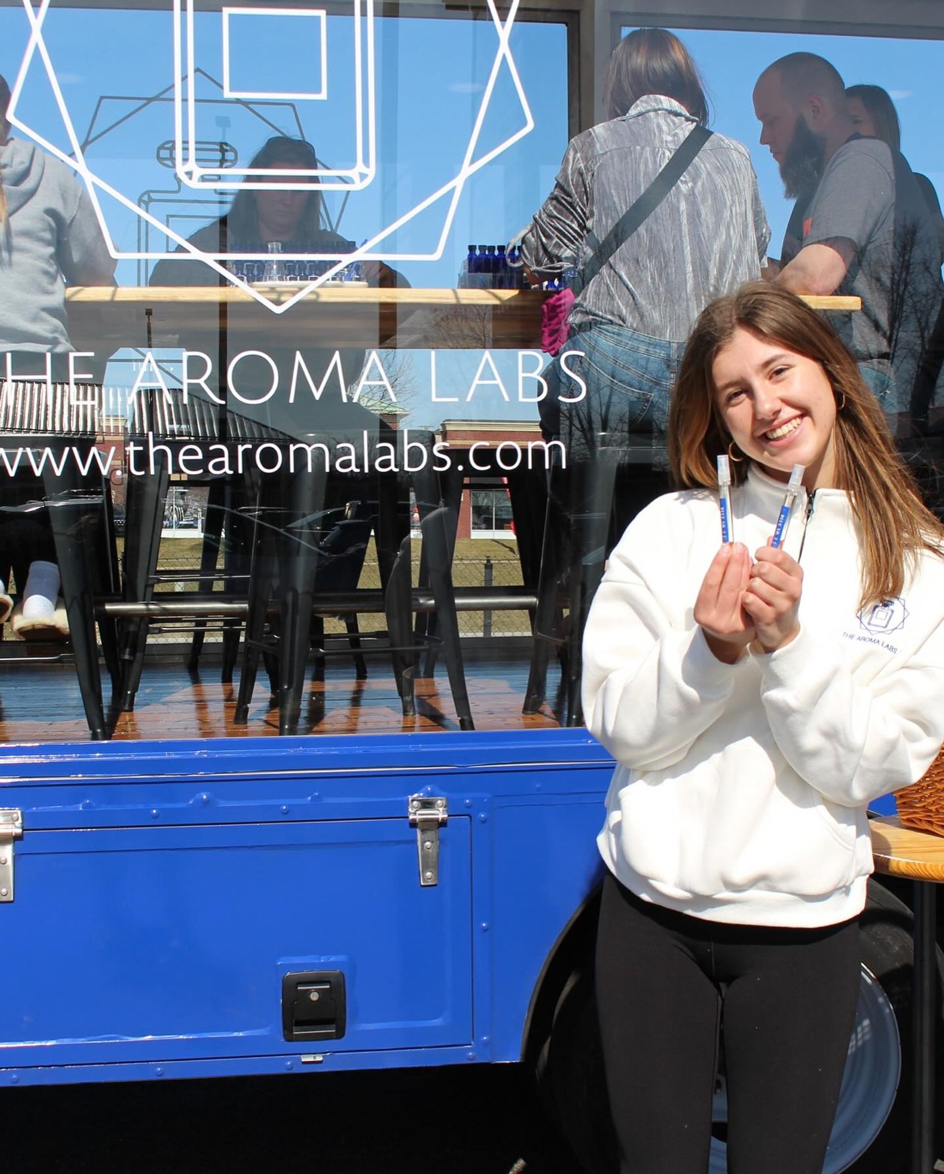 Smiling young woman holding two perfume sample vials outside a blue mobile fragrance pop-up truck with glass window seating and customers inside