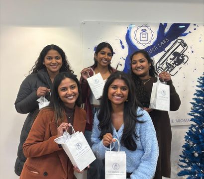 Five smiling women posing inside a bright shop, each holding a small white branded gift bag in front of a blue mural and a decorated blue holiday tree.
