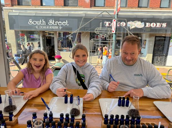 Smiling trio — two girls and an adult man — seated at a sunlit shop counter by a street window, blending scents from small blue bottles onto paper strips with coffee-bean sniff jars.