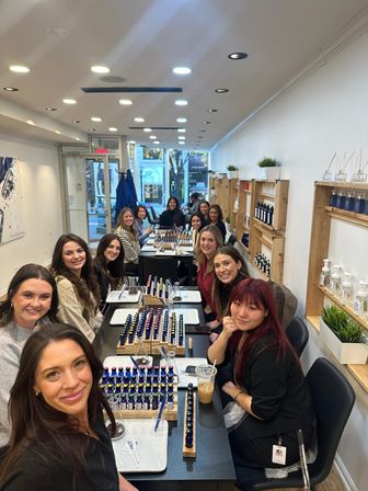 Smiling group of women at a bright downtown perfume-making workshop in a storefront studio, seated around a long table with rows of small blue fragrance bottles, droppers, mixing trays and wooden shelves displaying products and potted plants.