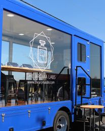 Bright blue mobile cafe truck with large glass window displaying a white geometric perfume-bottle graphic, wooden counter and black bar stools inside, outdoor step and round table under a clear blue sky.
