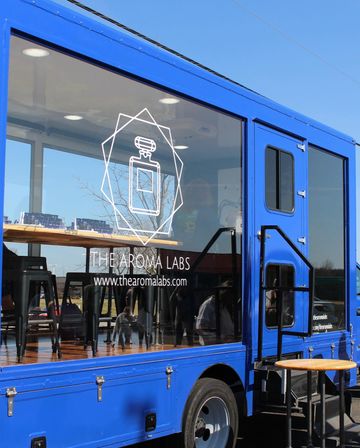 Bright blue mobile cafe truck with large glass window displaying a white geometric perfume-bottle graphic, wooden counter and black bar stools inside, outdoor step and round table under a clear blue sky.
