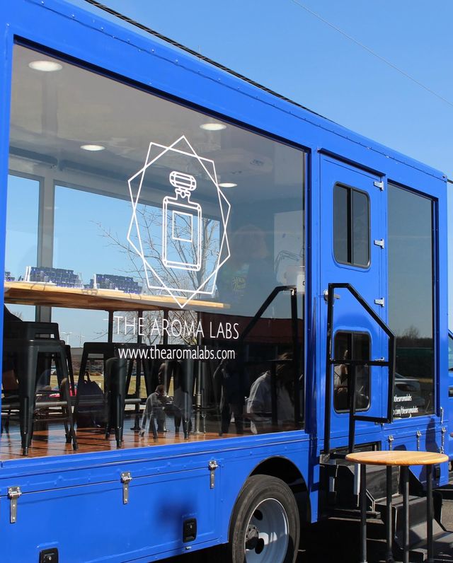 Bright blue mobile cafe truck with large glass window displaying a white geometric perfume-bottle graphic, wooden counter and black bar stools inside, outdoor step and round table under a clear blue sky.