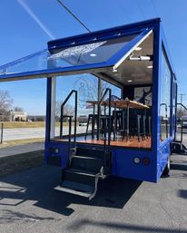 Blue mobile pop-up truck with fold-down glass side, wooden high-top table and black stools inside, metal steps up to the platform, parked in a sunny roadside lot