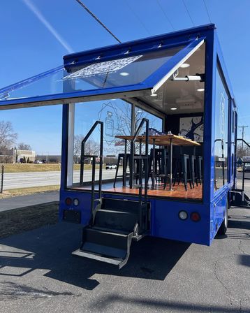 Blue mobile pop-up truck with fold-down glass side, wooden high-top table and black stools inside, metal steps up to the platform, parked in a sunny roadside lot