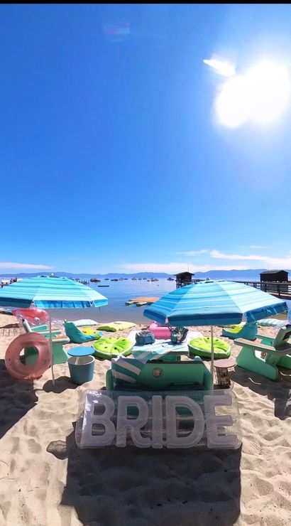 Sunlit lakeside beach with turquoise umbrellas, lounge chairs, colorful floats and large inflatable "BRIDE" letters by calm water and docks