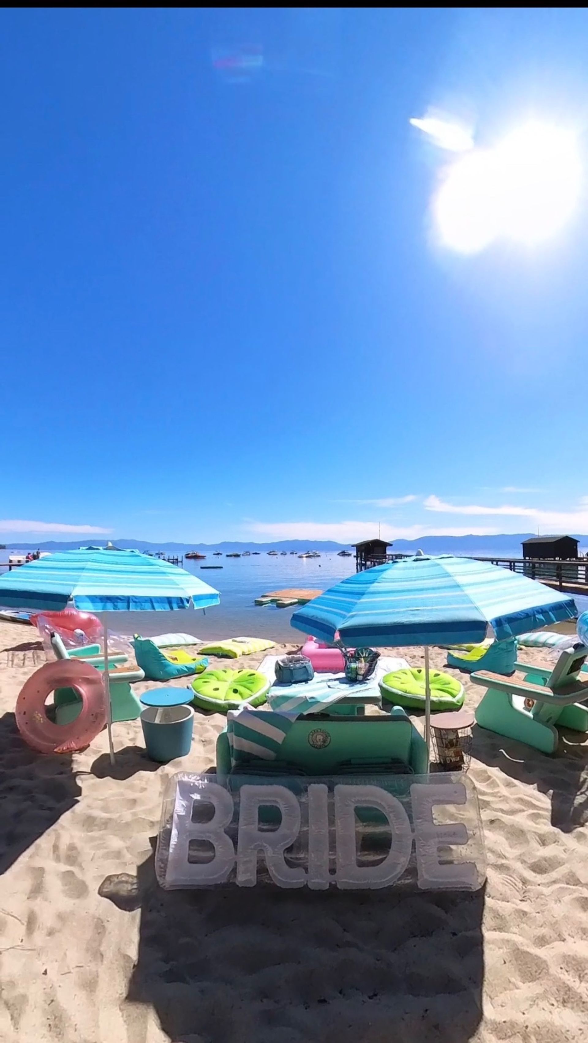 Sunlit lakeside beach with turquoise umbrellas, lounge chairs, colorful floats and large inflatable "BRIDE" letters by calm water and docks