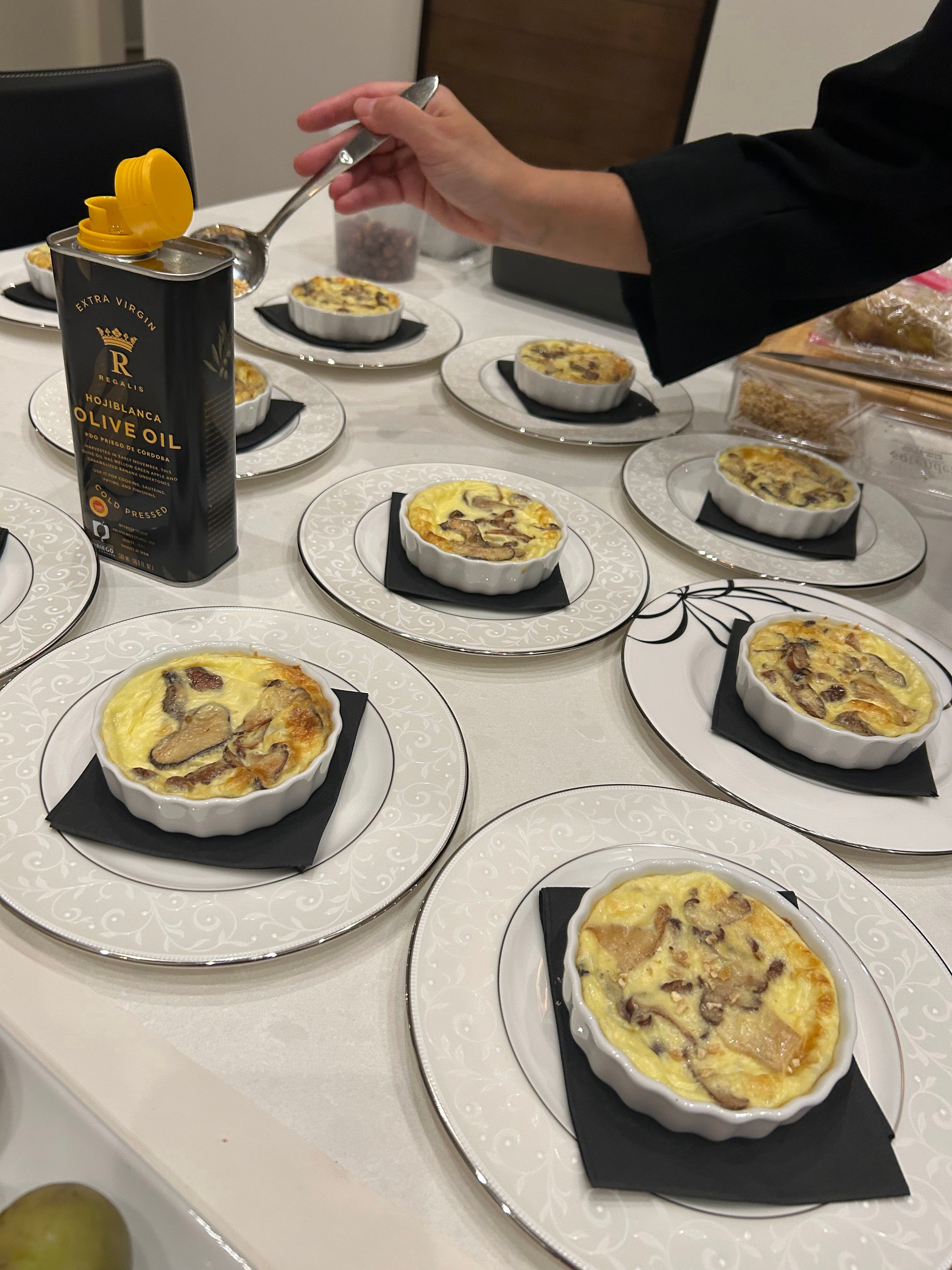 Plated mini mushroom and cheese quiches in white ramekins on decorative plates with black napkins, hand drizzling extra virgin olive oil — catered appetizer spread.