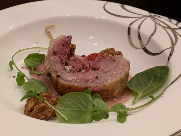 Plated rolled roast lamb with pink jus, cracked peppercorns, watercress and candied walnuts on a white decorative dinner plate — elegant fine-dining presentation.