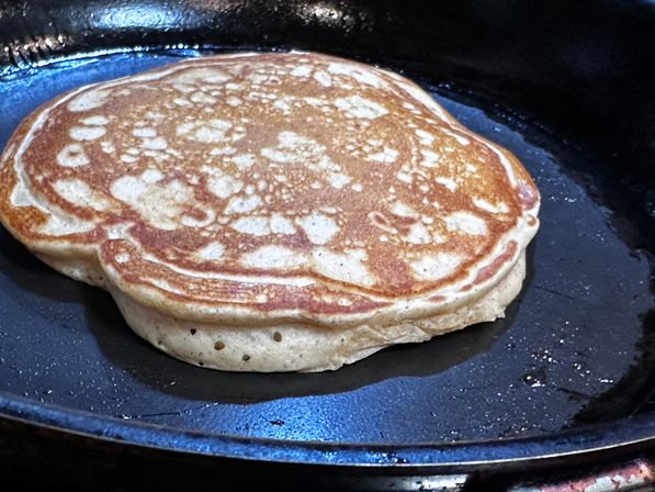 Close-up of a thick, golden-brown pancake cooking in a black cast-iron skillet — fluffy breakfast pancake on the stovetop.