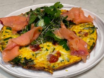 Close-up of golden frittata topped with smoked salmon ribbons, cherry tomatoes, fresh herbs and microgreens on a white plate — vibrant brunch dish.