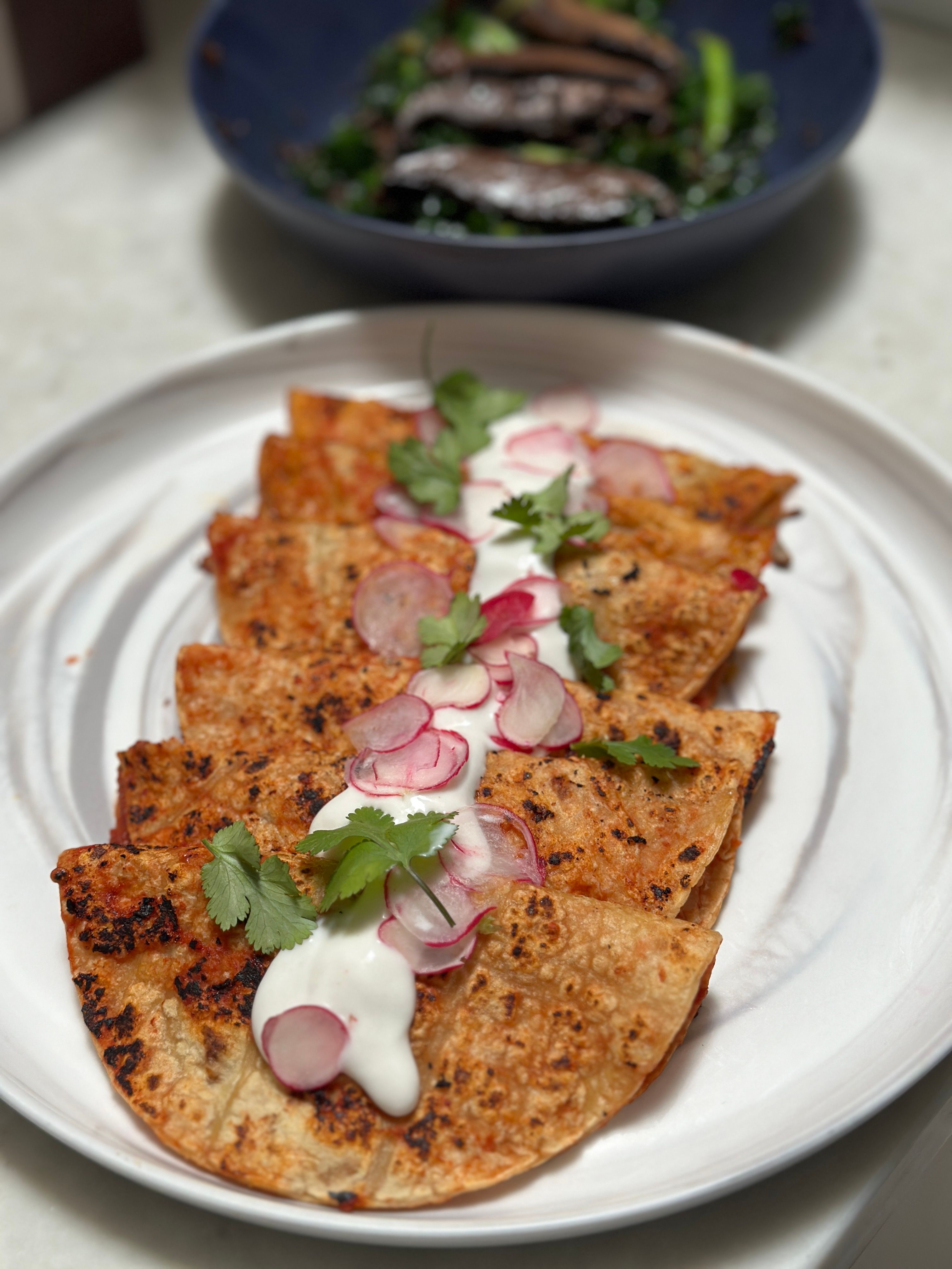 Crispy charred quesadillas arranged on a white plate, drizzled with crema and topped with thin radish slices and cilantro, with a bowl of greens blurred in the background.