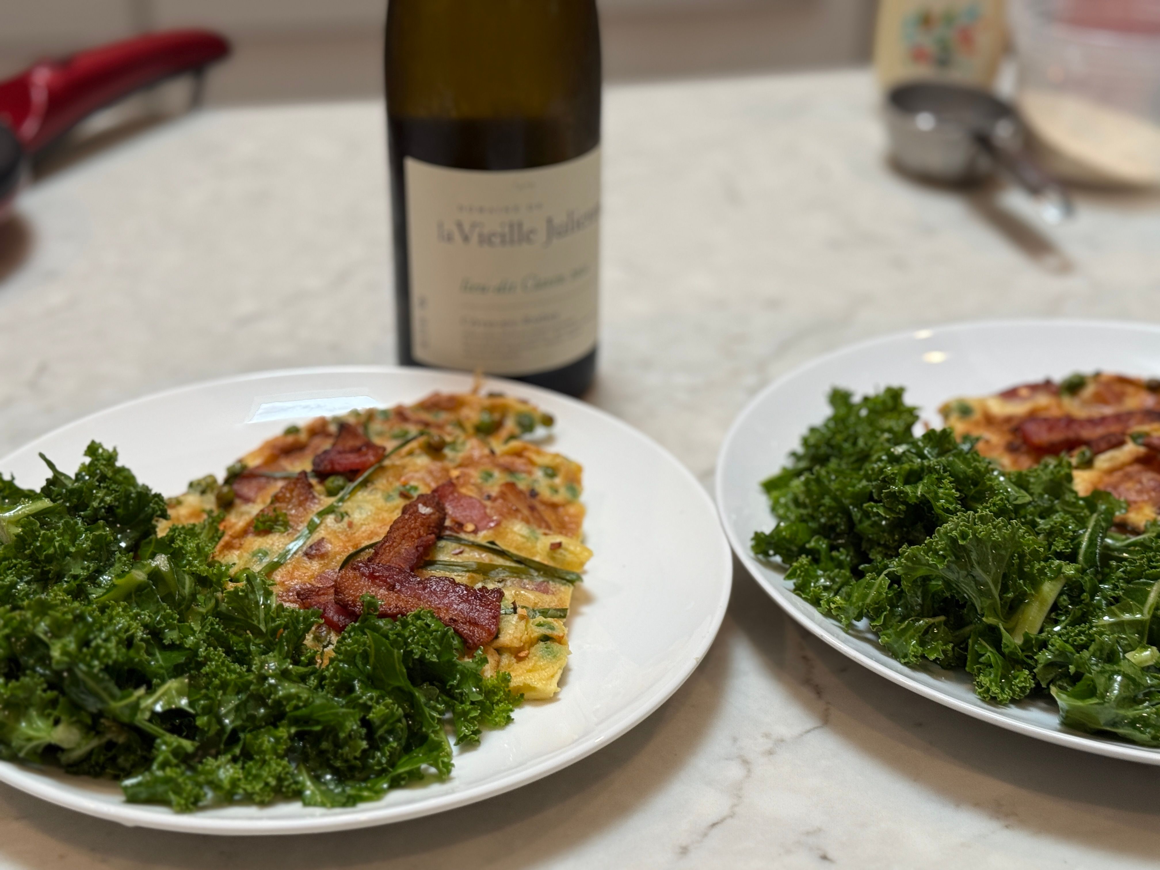 Two white plates with savory scallion egg pancakes topped with crispy bacon, served with a fresh kale salad on a marble countertop and a blurred bottle of white wine in the background