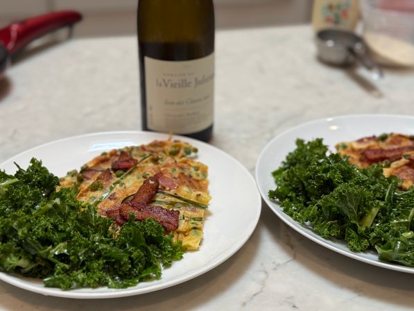 Two white plates with savory scallion egg pancakes topped with crispy bacon, served with a fresh kale salad on a marble countertop and a blurred bottle of white wine in the background