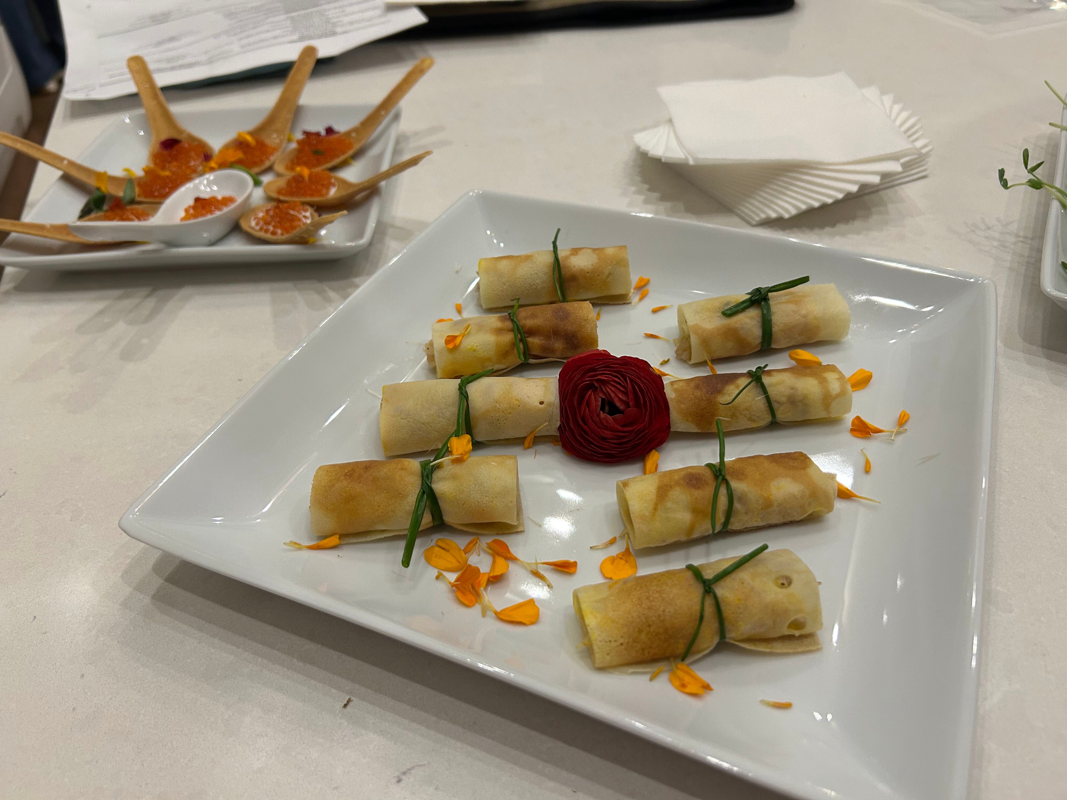 White square plate of eight bite-sized golden spring-roll appetizers tied with chive, scattered orange flower petals and a red rosette garnish, with wooden tasting spoons of orange roe on a tray in the background — elegant catering appetizer platter.