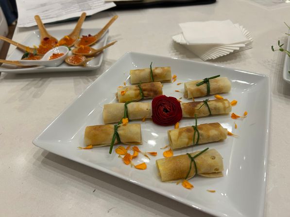 White square plate of eight bite-sized golden spring-roll appetizers tied with chive, scattered orange flower petals and a red rosette garnish, with wooden tasting spoons of orange roe on a tray in the background — elegant catering appetizer platter.