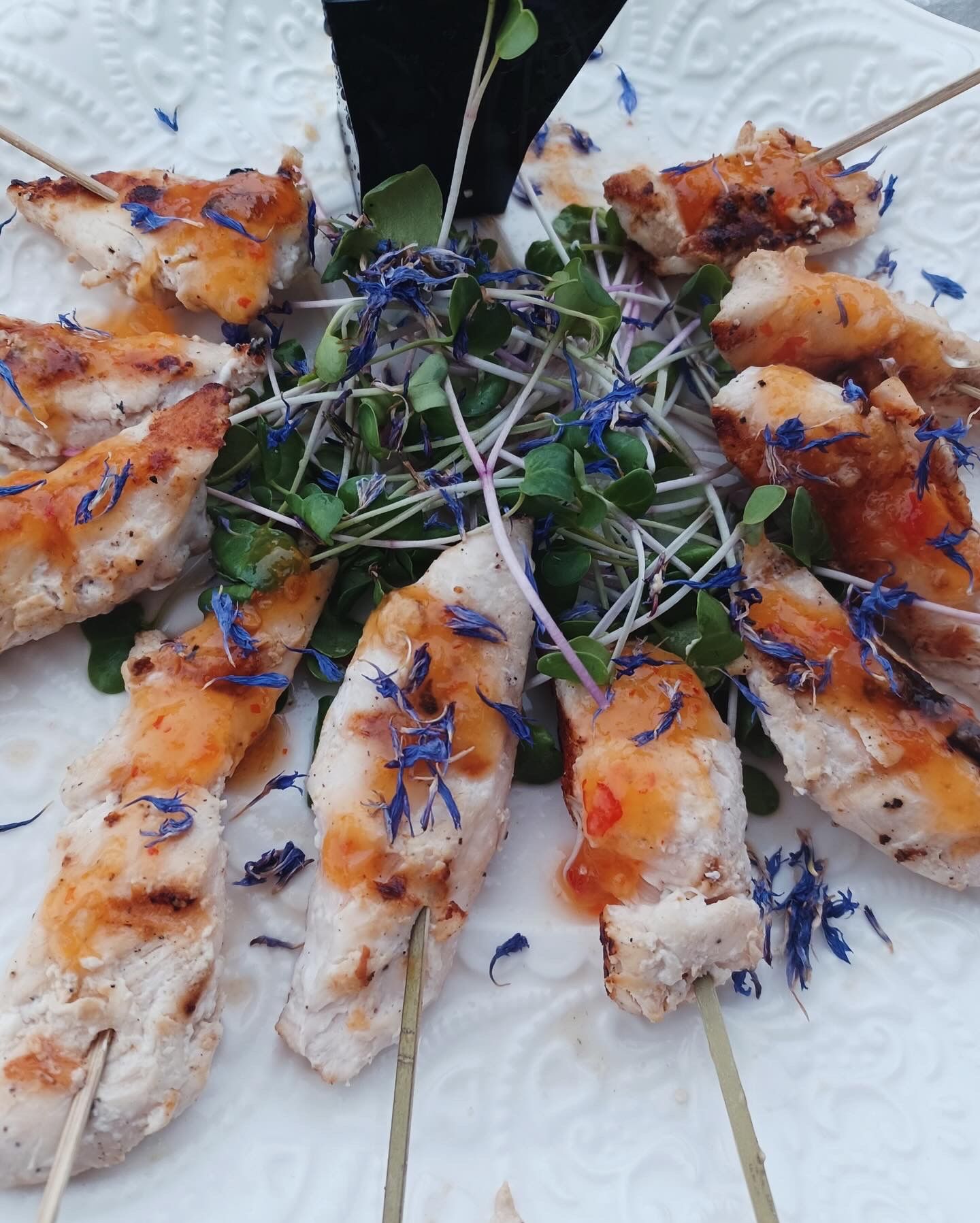 Grilled chicken skewers with sweet‑chili glaze and blue edible flower petals on a bed of microgreens, served on a white patterned plate — gourmet appetizer