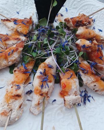 Grilled chicken skewers with sweet‑chili glaze and blue edible flower petals on a bed of microgreens, served on a white patterned plate — gourmet appetizer