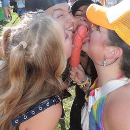 Four people at a sunny outdoor event in a grassy field playfully licking a novelty pink popsicle together