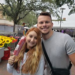 Two smiling adults at an outdoor street market holding a large wrapped red popsicle, with sunflowers, vendor tents and shady trees in the background