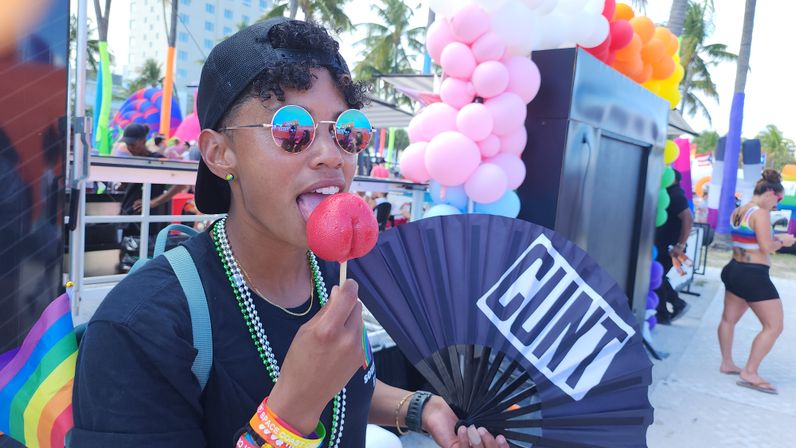 Festival-goer at a beachside Pride event wearing mirrored round sunglasses and a backwards cap, licking a shiny red candy apple on a stick and holding a black fan with bold white lettering, with rainbow balloons, flags and palm trees in the background.