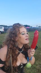 Curly-haired festivalgoer with blue shimmer eye makeup and layered necklaces playfully posing with a long red novelty ice pop in a grassy outdoor music festival field