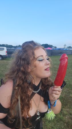 Curly-haired festivalgoer with blue shimmer eye makeup and layered necklaces playfully posing with a long red novelty ice pop in a grassy outdoor music festival field