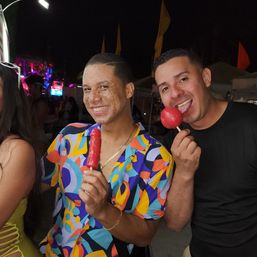 Two smiling men at a lively night market under festival lights and vendor tents — one in a colorful shirt holds a red popsicle while the other licks a red candy apple.