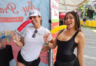 Two smiling women posing at an outdoor summer food festival vendor booth, each holding a large colorful frozen popsicle on a stick—one red, one yellow—with umbrellas and vendor tents in the background
