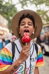 Person in a straw hat and rainbow-striped shirt playfully licking a glossy red candy apple on a stick at an outdoor street festival.