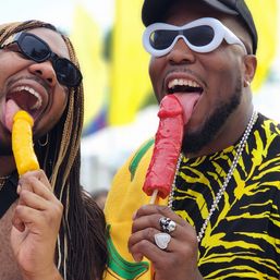 Two adults in sunglasses at an outdoor summer festival playfully licking colorful novelty popsicles (yellow and red) with bright flags and festive clothing in the background