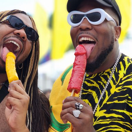 Two adults in sunglasses at an outdoor summer festival playfully licking colorful novelty popsicles (yellow and red) with bright flags and festive clothing in the background