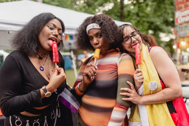Three people at an outdoor Pride festival enjoying colorful popsicles, posing under park tents in playful outfits with pins and small flags.